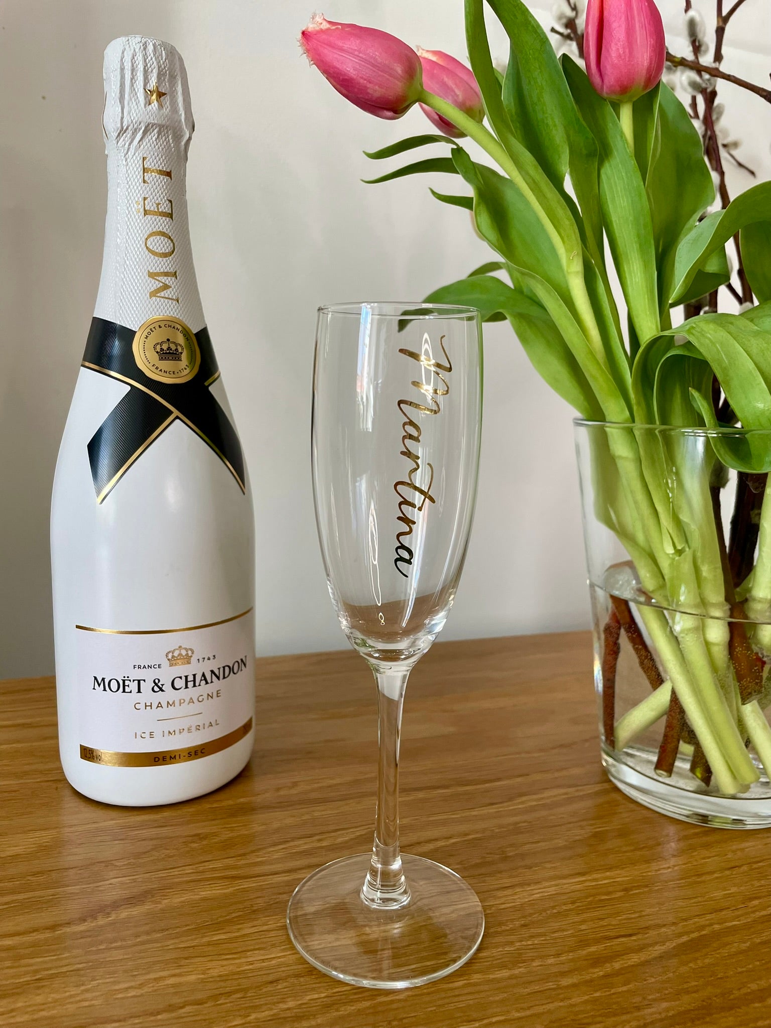 Personalisiertes Sektglas mit Namen - im Hintergrund eine Flasche Champage und Tulpen in Vase